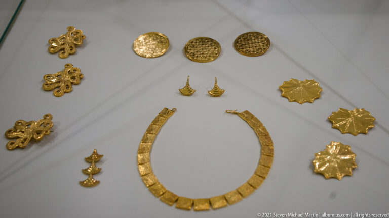 Gold necklace, circular discs, pendants, plaques, and openwork ornaments arranged together in a museum display case.