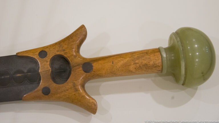 Wooden weapon hilt with dark metal fittings, circular inlays, and a rounded green stone pommel.