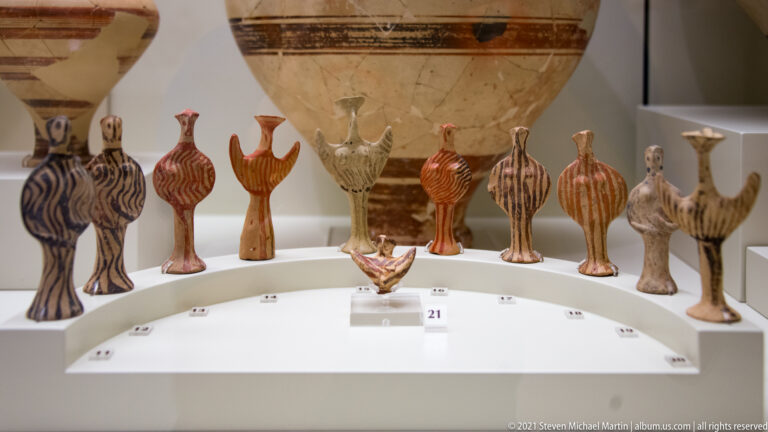 Group of painted Mycenaean terracotta figurines with striped bodies and raised or projecting arms in a museum case.