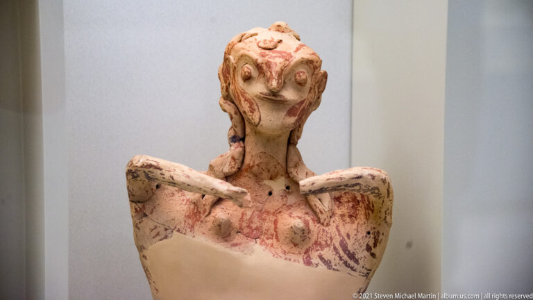Painted terracotta female figurine with raised arms, modeled face, and surviving red pigment on the torso.