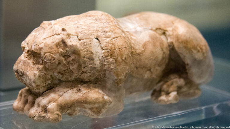 Small pale stone sculpture of a reclining lion-like animal with head lowered toward its forepaws.
