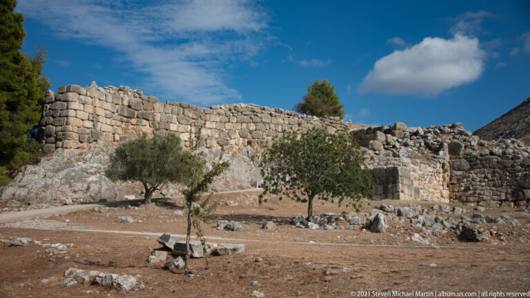 Massive stone fortification wall built over exposed bedrock with olive trees and archaeological remains in front.
