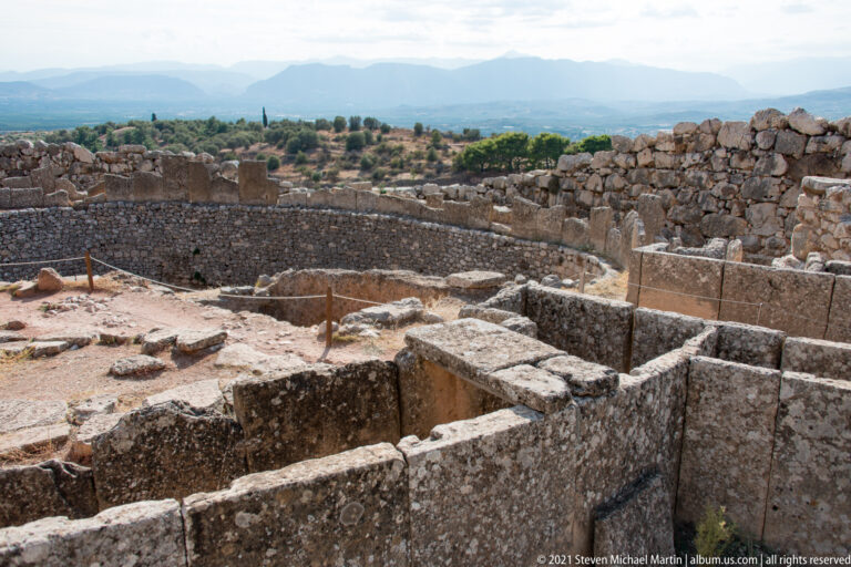 Low stone foundations and rectangular rooms on the upper citadel with the Argolid landscape beyond.