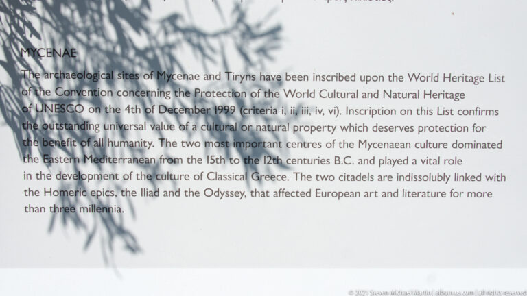 Close view of English UNESCO inscription text for Mycenae and Tiryns with leaf shadows across the panel.