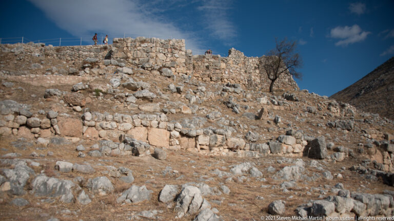Terraced stone walls and foundations crossing a dry rocky slope within the Mycenaean citadel.