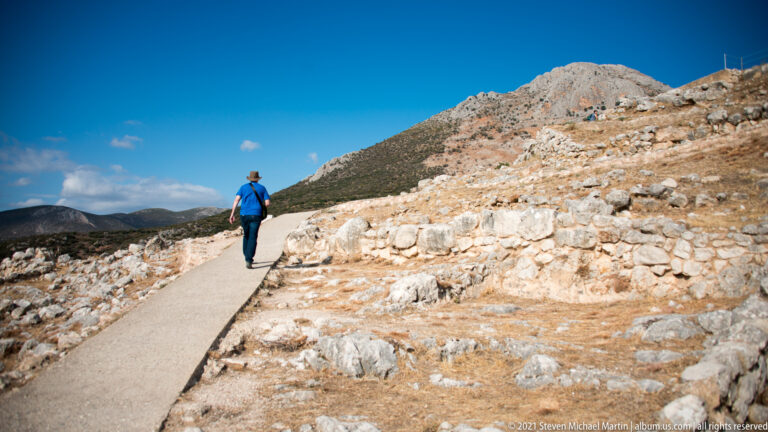 Visitor walking uphill on a paved path between low stone ruins under a bright blue sky.