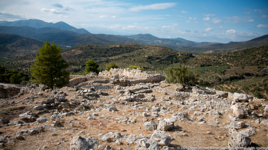 Stone ruins and a lone pine on the upper citadel overlooking the broad hilly Argolid landscape.