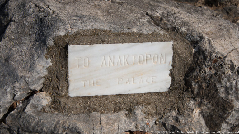 Rectangular stone wayfinding marker set into rock, inscribed in Greek and English with 'THE PALACE.'