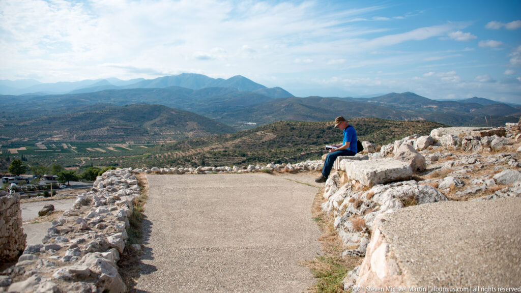 Visitor seated beside a curving upper-citadel path with stone ruins and distant mountain landscape beyond.