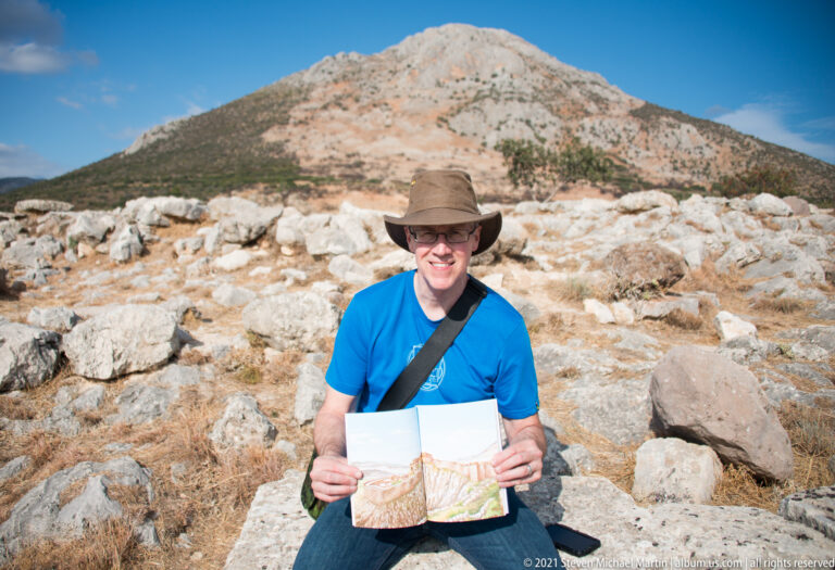 Visitor in blue shirt and hat holding an open site map among rocky ruins with a mountain behind.