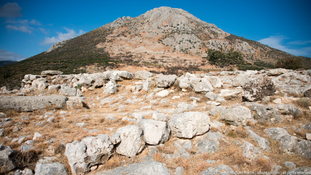 Low stone archaeological remains in dry terrain beneath a steep pale mountain and deep blue sky.