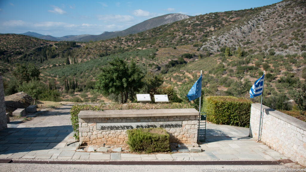 Paved approach at Ancient Mycenae with Greek and blue flags, low stone walls, hedges, and mountain slopes.