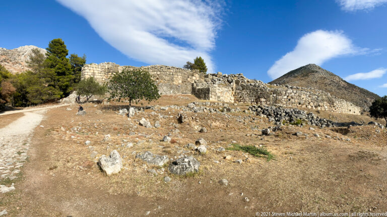 Stone walls and archaeological terraces spread across a dry slope beneath a steep rocky mountain.