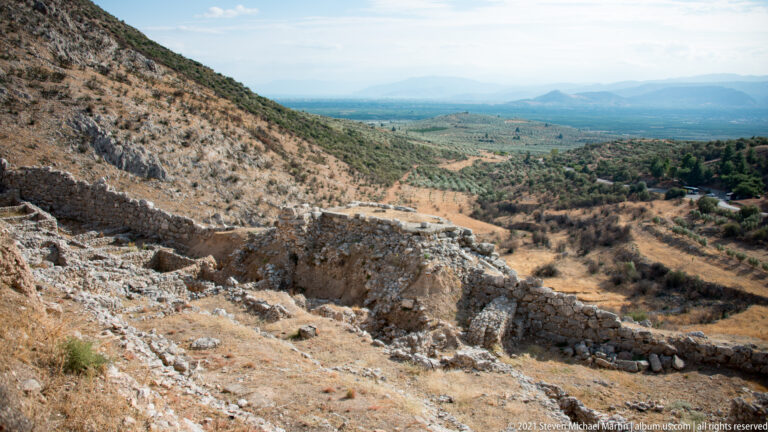 Stone fortification wall descending across a dry hillside with the Argolid plain and mountains beyond.