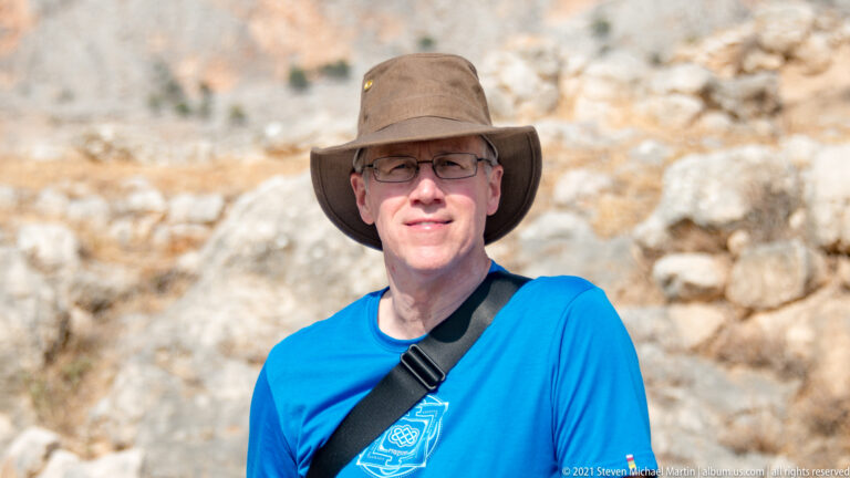 Visitor wearing glasses, a brown sun hat, and blue shirt standing before blurred stone ruins.