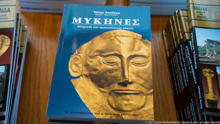 Greek-language book titled “ΜΥΚΗΝΕΣ” with a gold mask on the cover, displayed among archaeological publications.