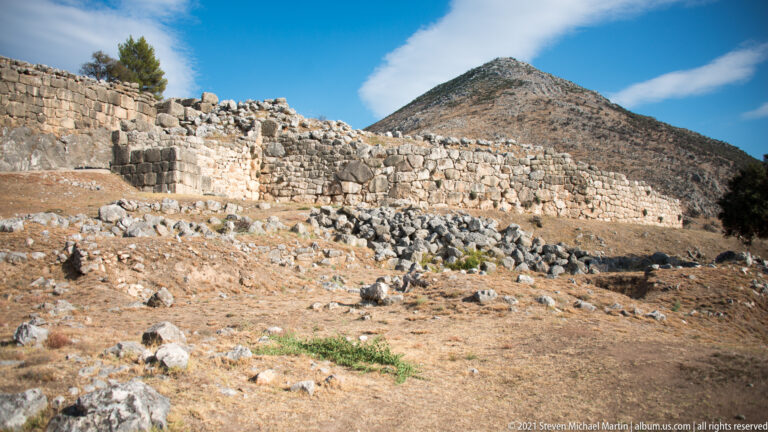 Long stone fortification wall stretching across dry ruins beneath a steep mountain and blue sky.