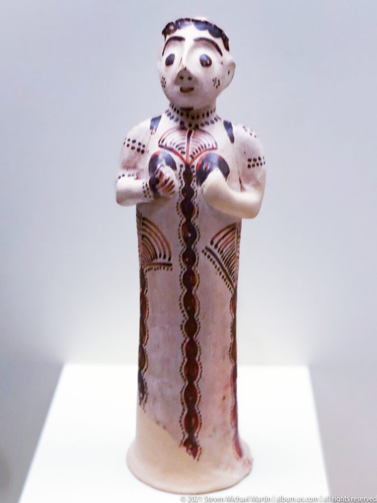 Standing painted terracotta female figurine with modeled face, folded arms, and red-brown linear decoration.