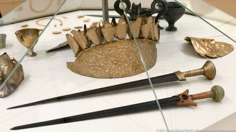 Two long swords displayed with gold ornaments, vessels, fittings, and other Mycenaean grave goods in a museum case.