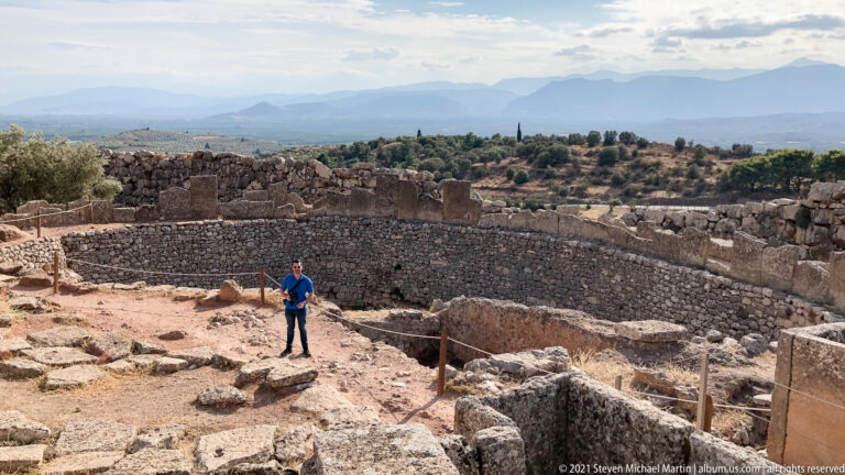 Visitor beside Grave Circle A at Mycenae, with upright grave stelae, circular enclosure wall, and the Argolid landscape beyond.