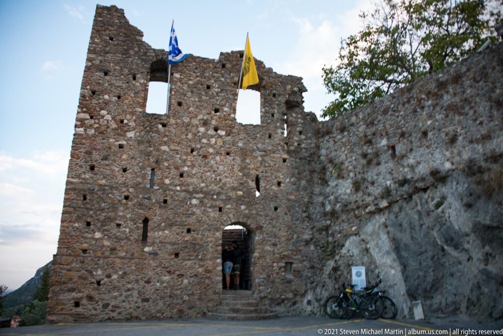 Stone gateway at Mystras with Greek and Byzantine flags above its ruined parapet and arched entrance.