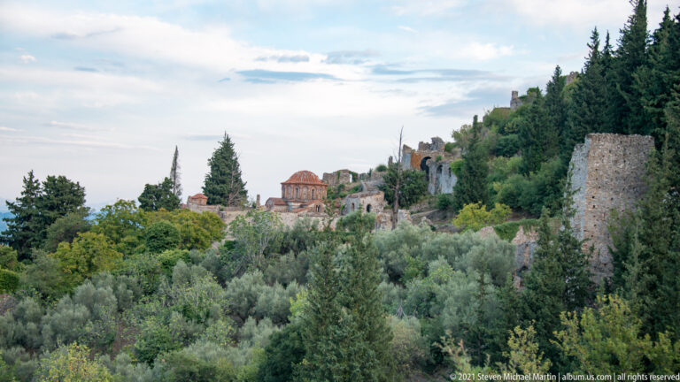 Domed Byzantine church, likely Hagia Sophia, among cypress trees and ruins on the Mystras hillside.