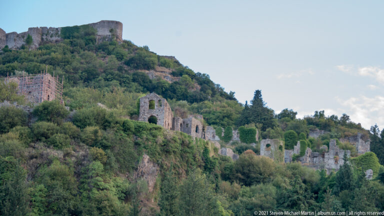 Roofless stone ruins with arched openings extend across the Mystras slope below the fortress.