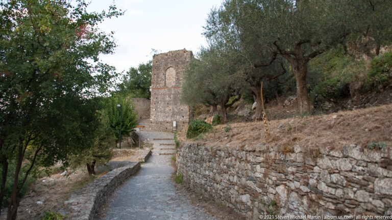 Stone-paved path climbs between olive trees and walls toward a tall masonry ruin at Mystras.
