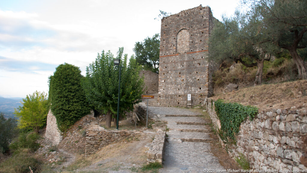 Stepped stone lane leading toward a tall masonry structure on the hillside at Mystras, with olive trees and valley views.
