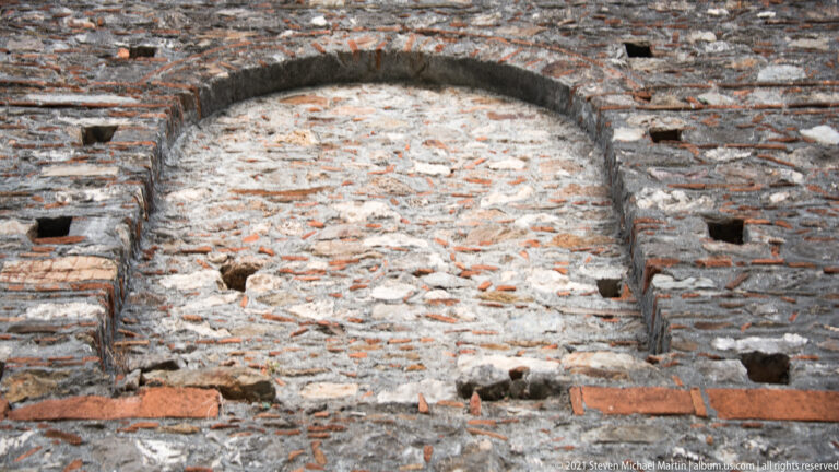 Large brick-framed arch at Mystras filled with rubble masonry, with horizontal brick bands and small square wall sockets.