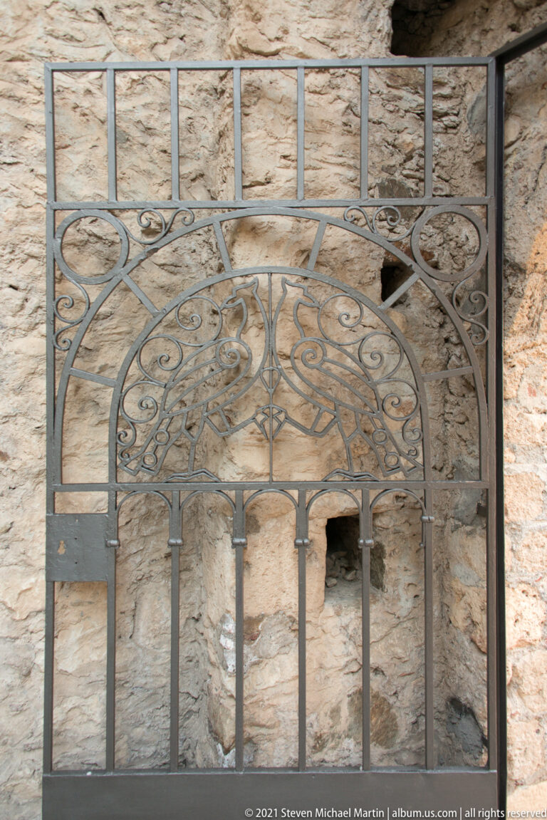 Ornamental metal grille with scrollwork mounted before a small recessed opening in a stone wall at Mystras.