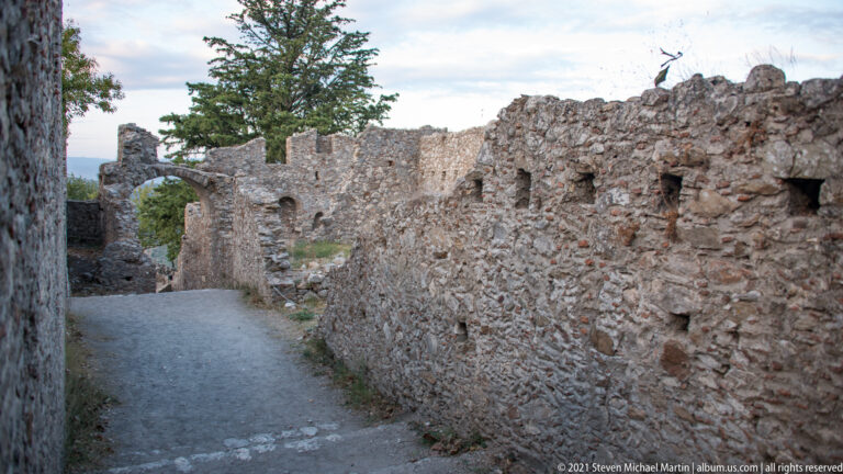 Narrow path through ruined masonry walls at Mystras, with broken wall tops and a glimpse of the valley beyond.