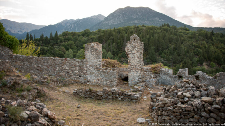 Low stone ruins and isolated masonry piers on the Mystras hillside with Mount Taygetos rising behind forested slopes.