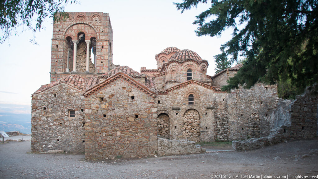 Byzantine church at Mystras with tiled domes, patterned brickwork, narrow arched windows, and a tall open bell tower.