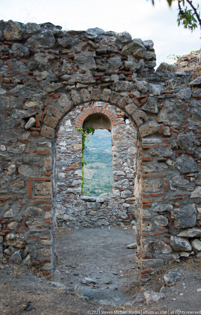 Semicircular brick-framed doorway in a ruined Mystras wall opening toward the green valley beyond.