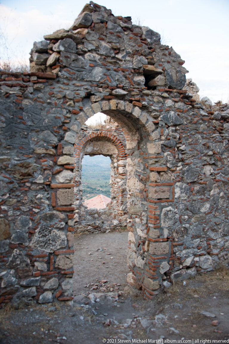 Brick-framed semicircular doorway in a ruined stone wall at Mystras, with another arch and distant landscape visible beyond.