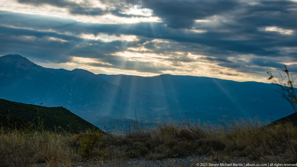 Sunbeams stream through dark clouds over layered mountain ridges in the landscape surrounding Mystras.