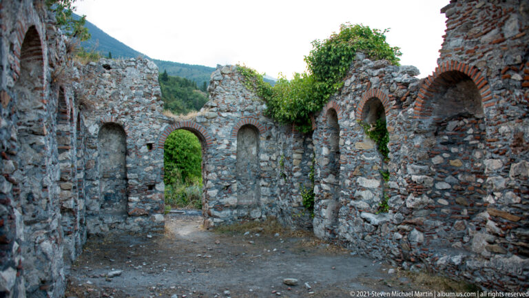 Roofless chamber at Mystras with brick-framed arched recesses, rough stone walls, and a central doorway opening into greenery.