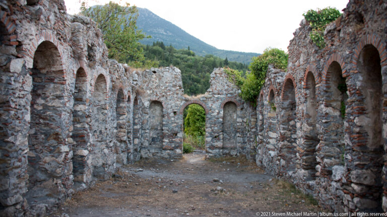 Long roofless chamber at Mystras with repeated arched recesses, a central doorway, and Mount Taygetos visible beyond.