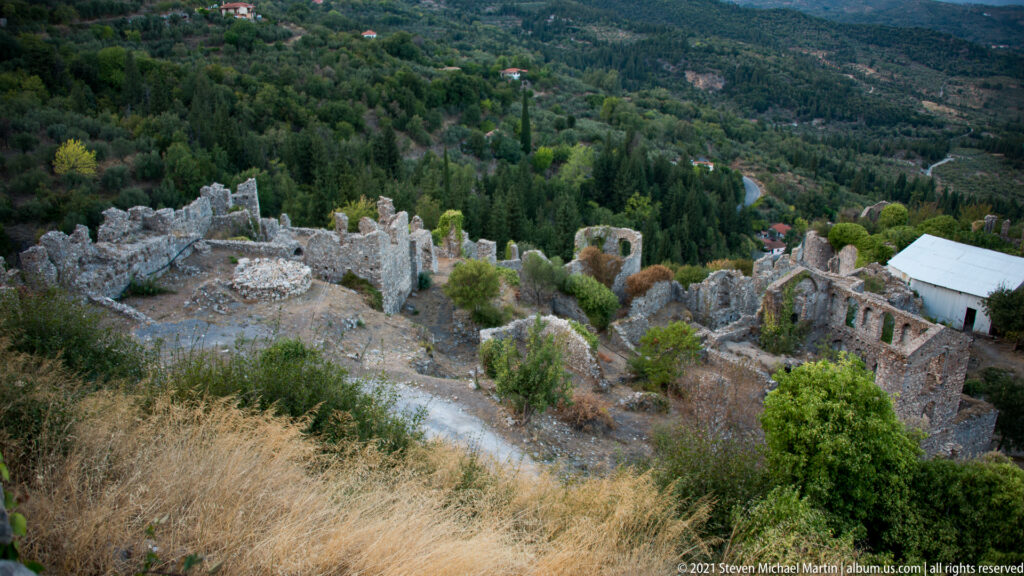 Elevated view over roofless stone ruins and vegetation on a lower terrace of Mystras, with a modern protective roof among the remains.