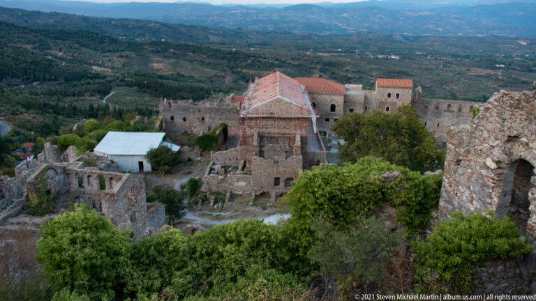 Elevated view of a red-tiled Byzantine church complex at Mystras with scaffolding, surrounding ruins, trees, and valley beyond.