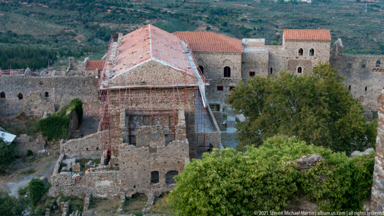 Scaffolded Byzantine church at Mystras with red-tiled roof, adjoining masonry buildings, trees, and fortified walls.