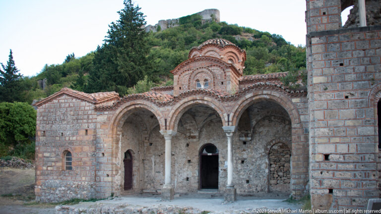 Three-arched Byzantine church portico at Mystras with marble columns, brick-and-stone arches, tiled dome, and bell-tower wall.
