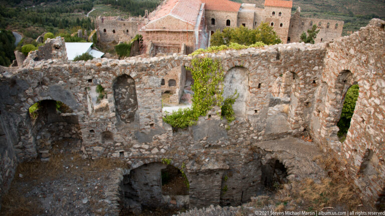 Roofless multi-level stone ruins at Mystras with arched openings and a conserved church complex visible beyond.