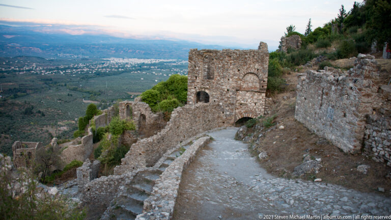 Paved path descending between ruined walls at Mystras toward a square masonry structure, with the broad valley below.