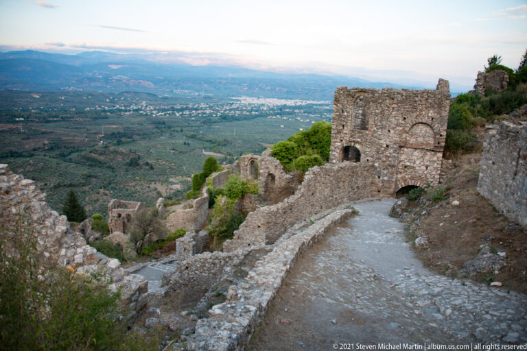 Descending path and ruined masonry at Mystras overlooking the broad plain and distant settlement below.
