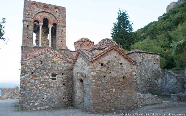 Exterior of St. Sophia at Mystras with stone walls, tiled roofs, domed upper structure, and tall open bell tower.