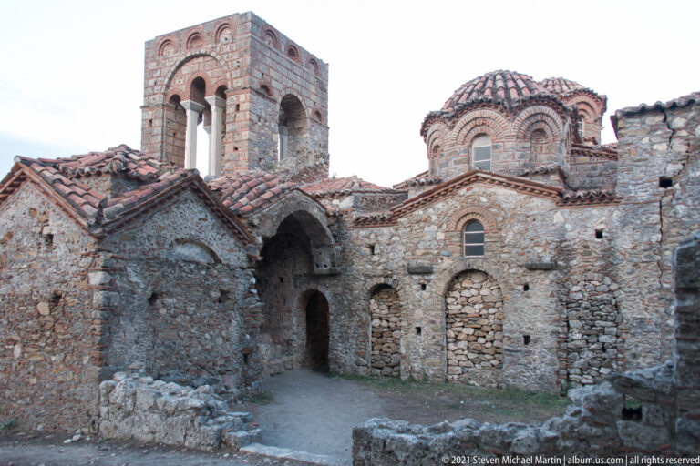 Courtyard view of St. Sophia at Mystras with bell tower, tiled roofs, dome, stone walls, and arched doorway.
