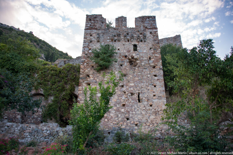 Tall rectangular stone tower with narrow openings and broken crenellations among vegetation at Mystras.