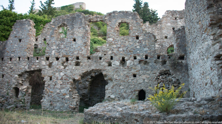 Tall roofless masonry ruin at Mystras with arched openings, rows of beam sockets, and vegetation growing through the structure.